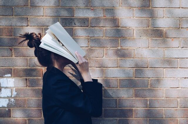 A person holds a book in front of their face while standing against a brick wall.