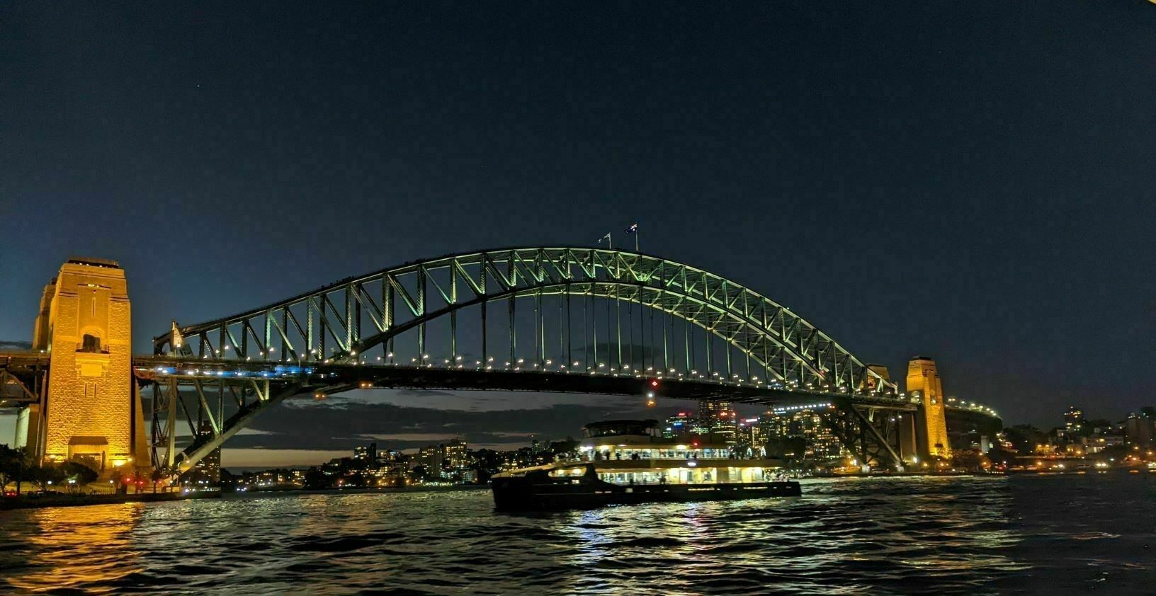 Sydney Harbour Bridge at night, with a lit-up ferry passing underneath and city lights in the background.