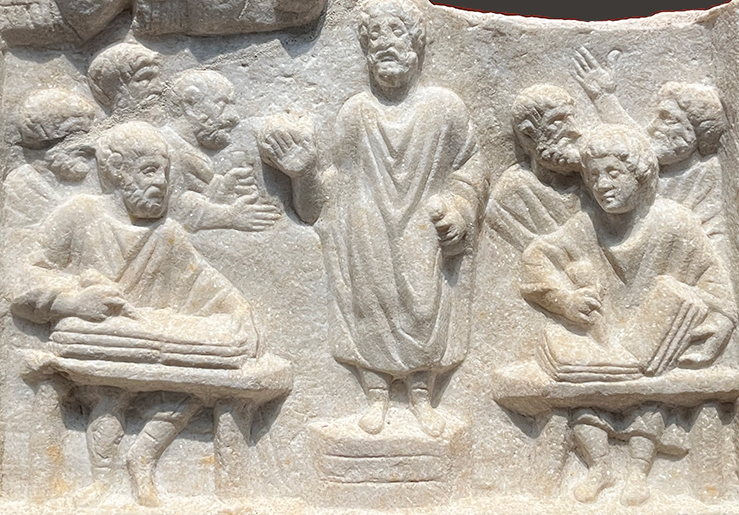 Auto-generated description: A group of figures in ancient attire is depicted in a carved stone relief, with some seated and writing as a central figure gestures.