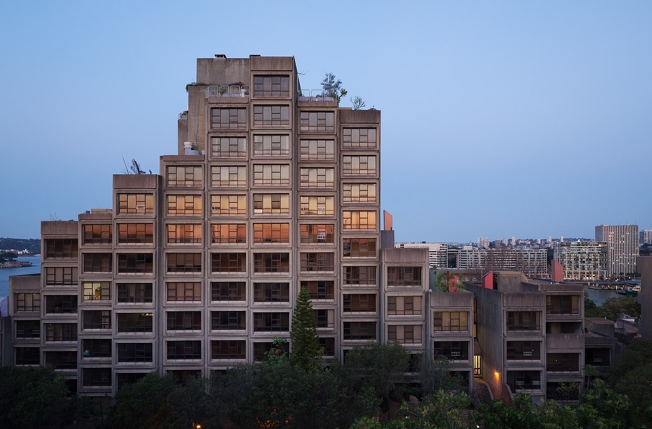 The distinctive, block-style concrete Sirius Building is set against an urban Sydney backdrop with a twilight sky.