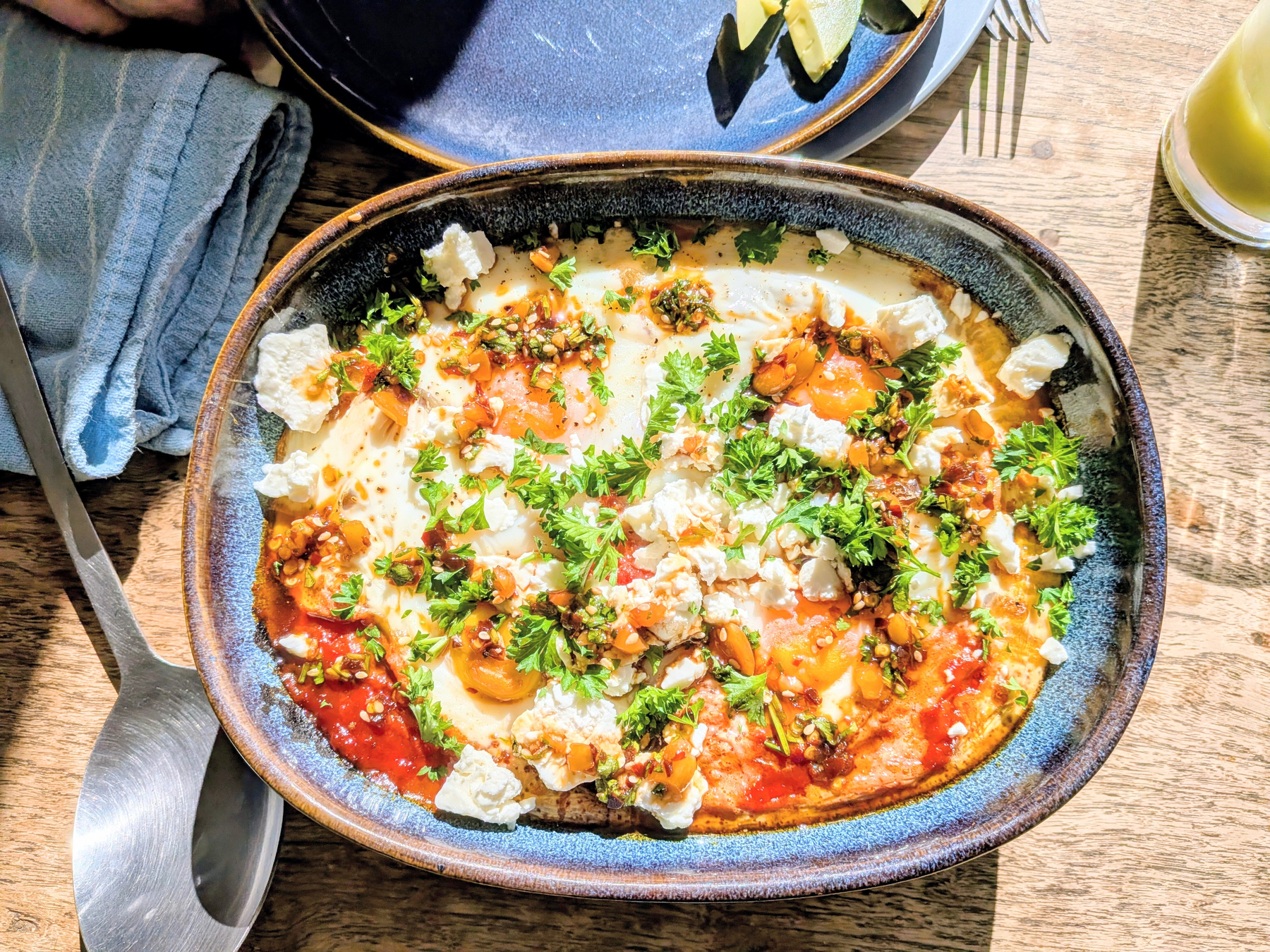 A dish of shakshuka - baked eggs topped with herbs, crumbled cheese, and a tomato-based sauce is served in an oval ceramic bowl on a wooden table.