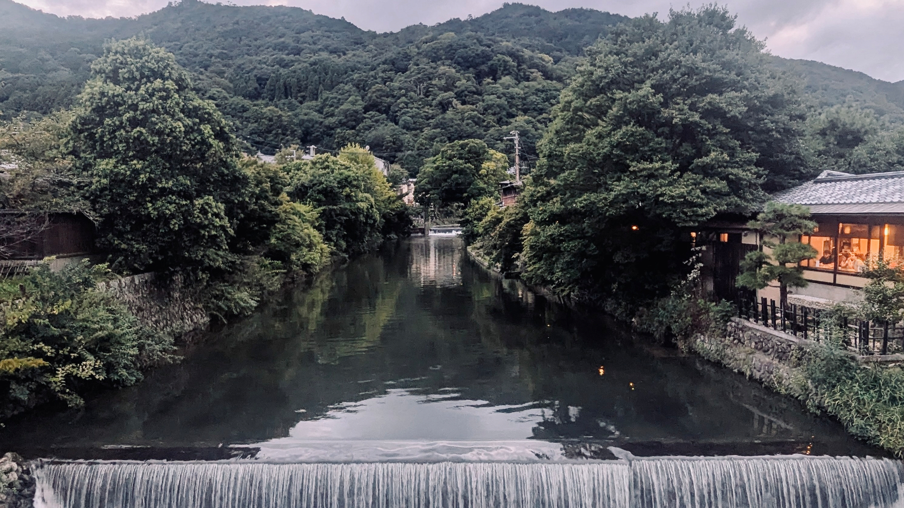 A serene river channel flows between two small weirs surrounded by lush greenery and mountains, with buildings on the right.