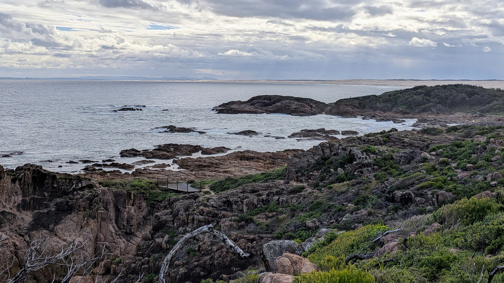 A rocky coastline with patches of greenery meets a calm sea under a cloudy sky. A long beach stretches out into the distance.