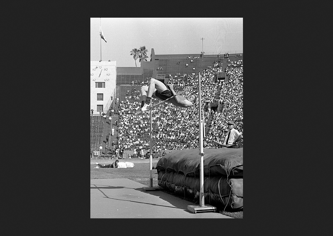 High jumper Dick Fosbury clearing the bar during 1968 Olympic trials at Los Angeles Memorial Coliseum, which is filled with spectators.