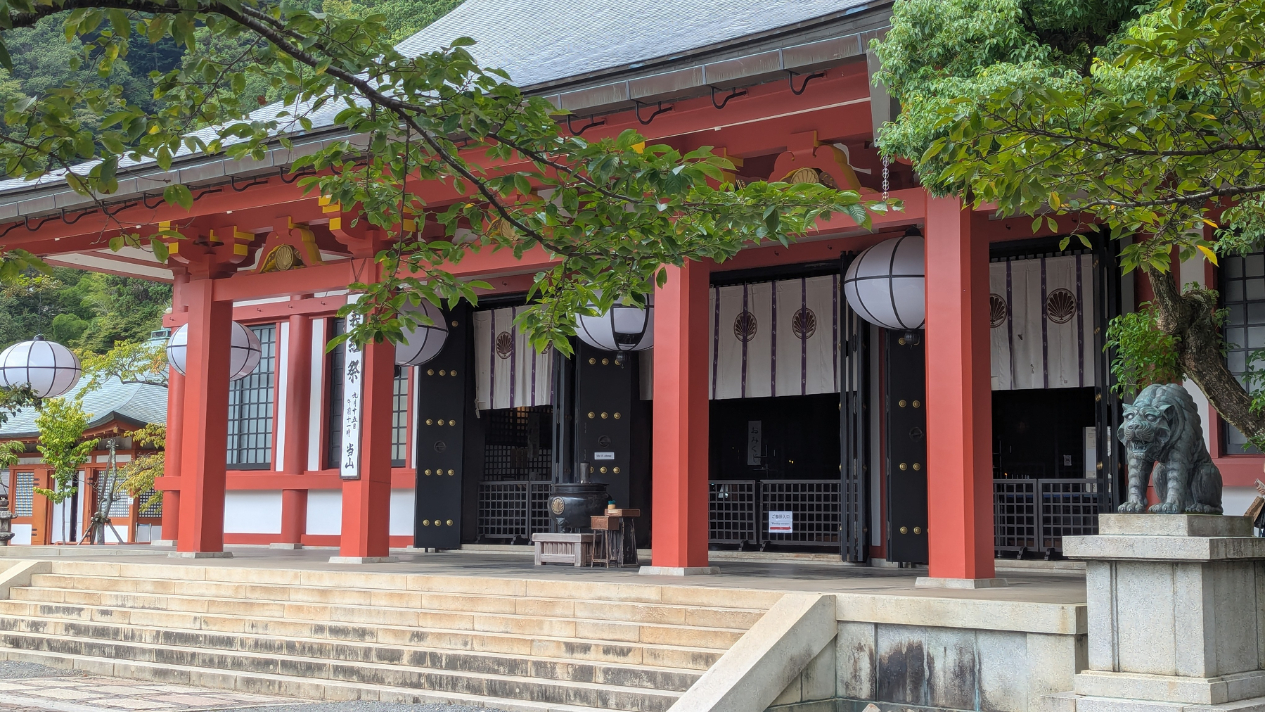 The main shrine at Kurama-dera with red columns, hanging lanterns, and a stone lion statue at the entrance, surrounded by lush greenery.