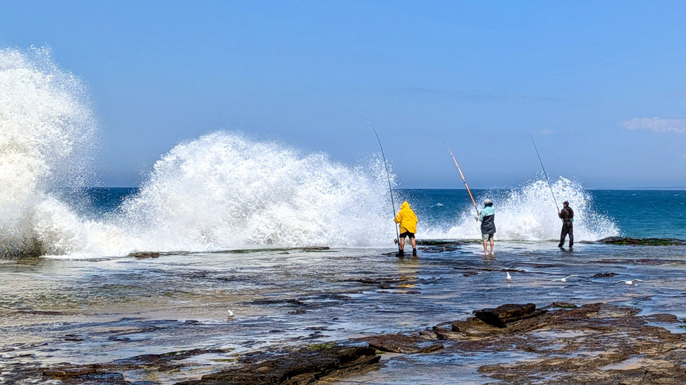 Three people are fishing on a rocky shore as large waves crash dramatically in front of them.