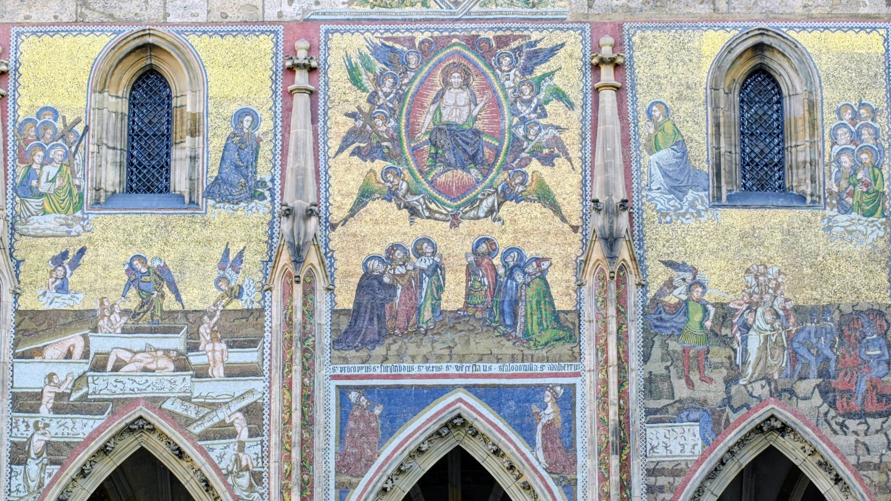 A detailed mosaic artwork on a cathedral facade depicts various religious scenes and figures surrounding arched windows.