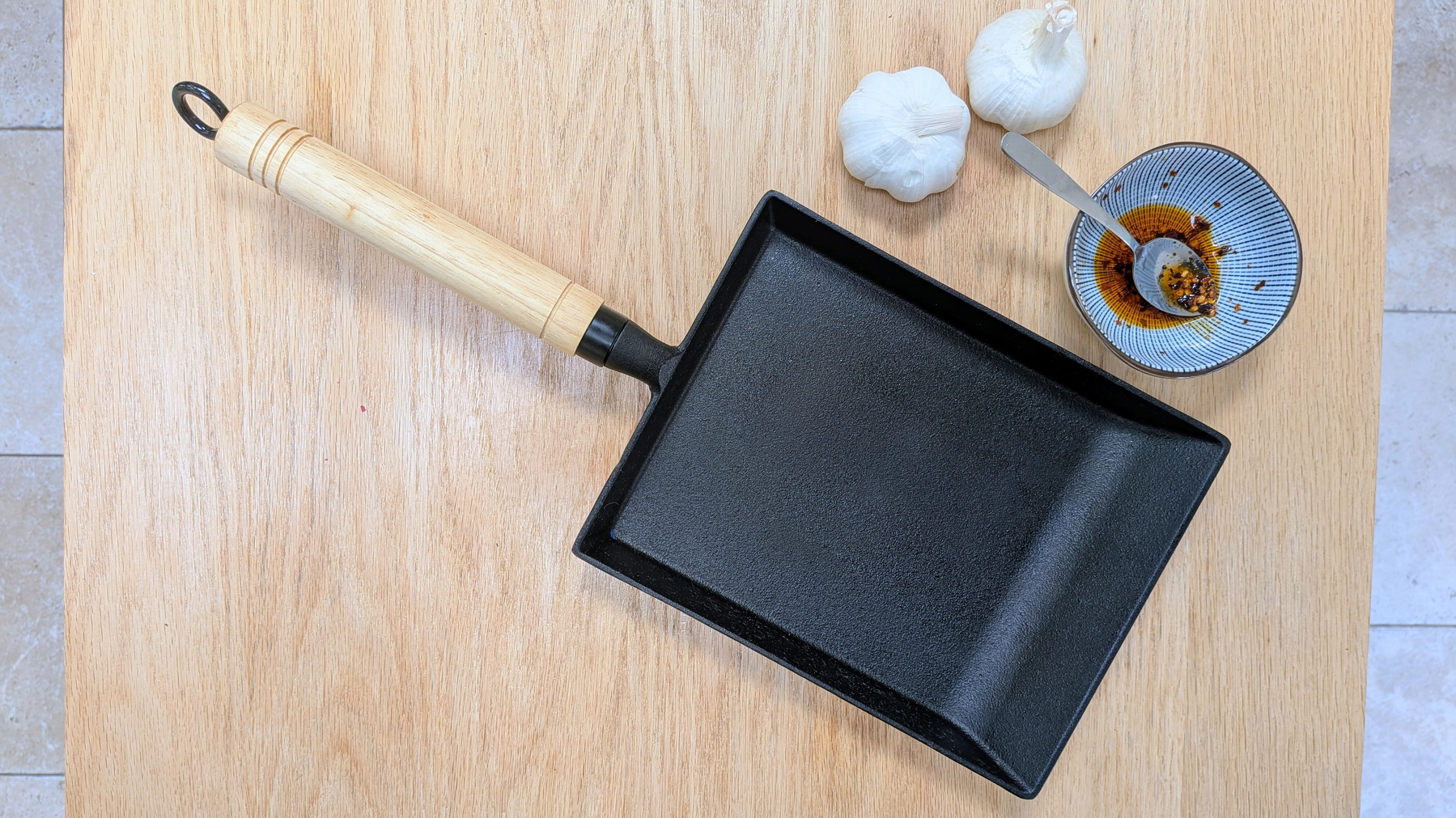 A rectangular black frying pan, or makiyakinabe, with a wooden handle is placed on a wooden surface next to two whole garlic bulbs and a small bowl containing chili flakes with a metal spoon.