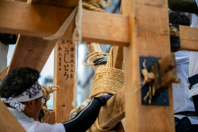 People are assembling a wooden structure using ropes, with some Japanese text visible on the wood.