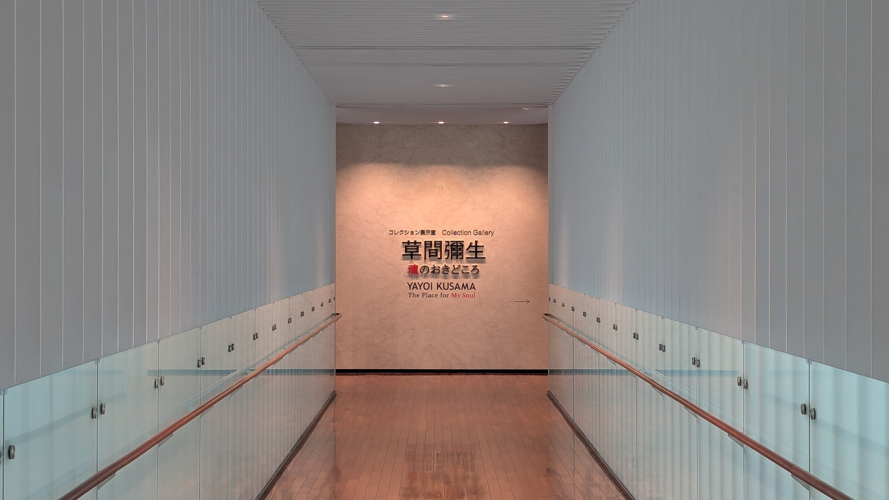 A narrow modern hallway with glass railings leads to a wall displaying text about Yayoi Kusama's exhibition in both Japanese and English.