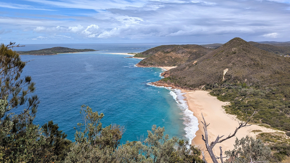 A scenic coastline featuring a sandy beach, an island, clear blue water, and hills covered with vegetation under a partly cloudy sky.