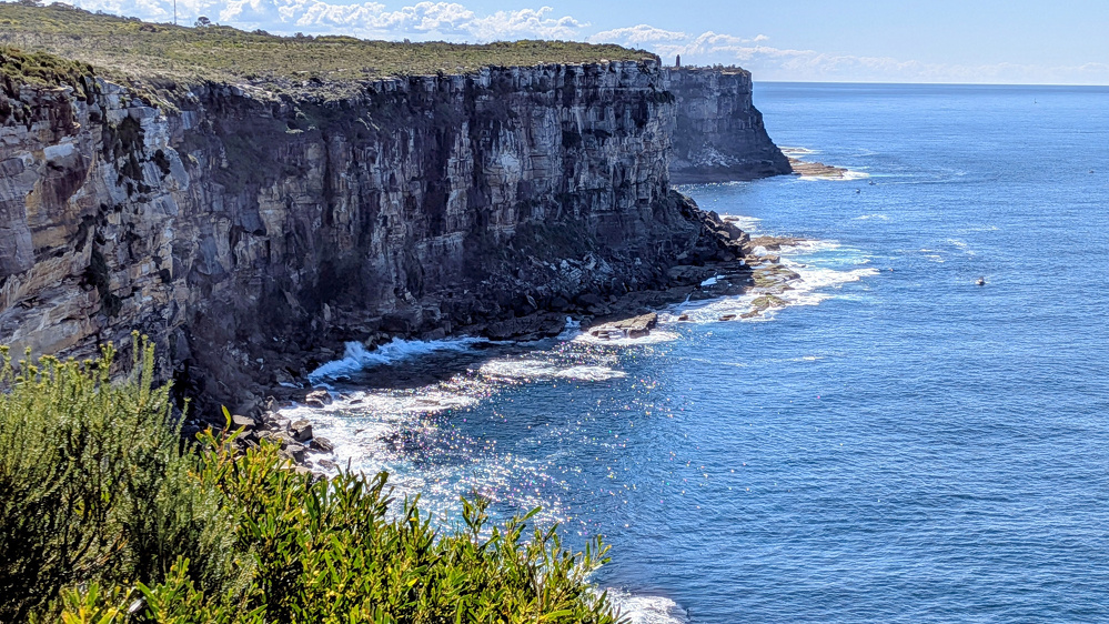 A rocky cliffside at North Head, Sydney, overlooks the ocean with waves gently crashing against the shore and some greenery in the foreground.