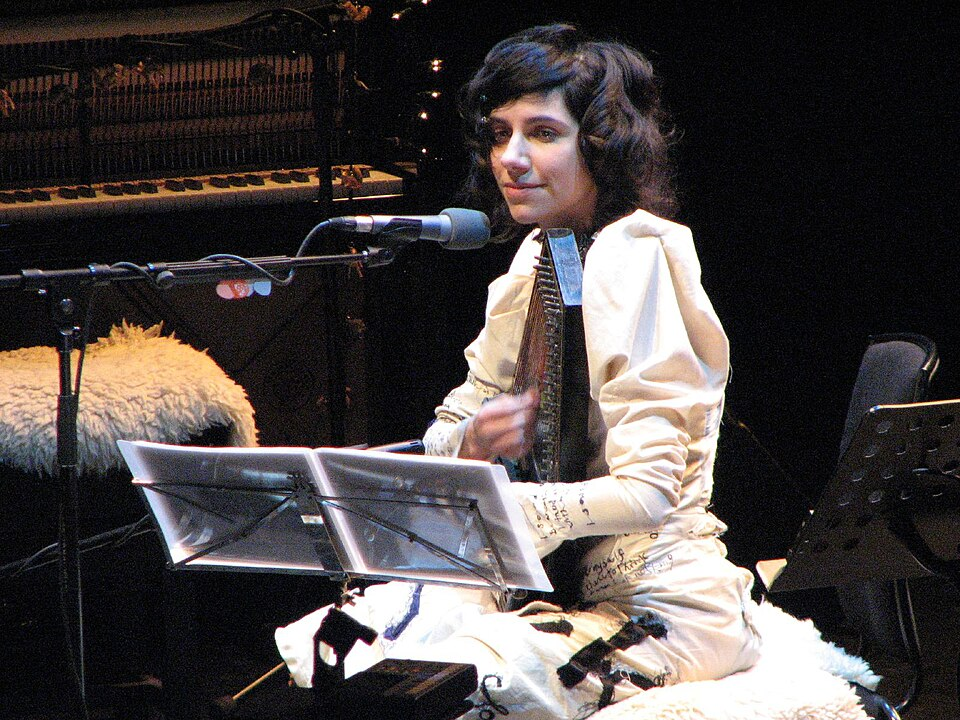 PJ Harvey is performing on stage, playing an autoharp and sitting behind a microphone with sheet music in front of her. In the background there's an upright piano with the wires visible.