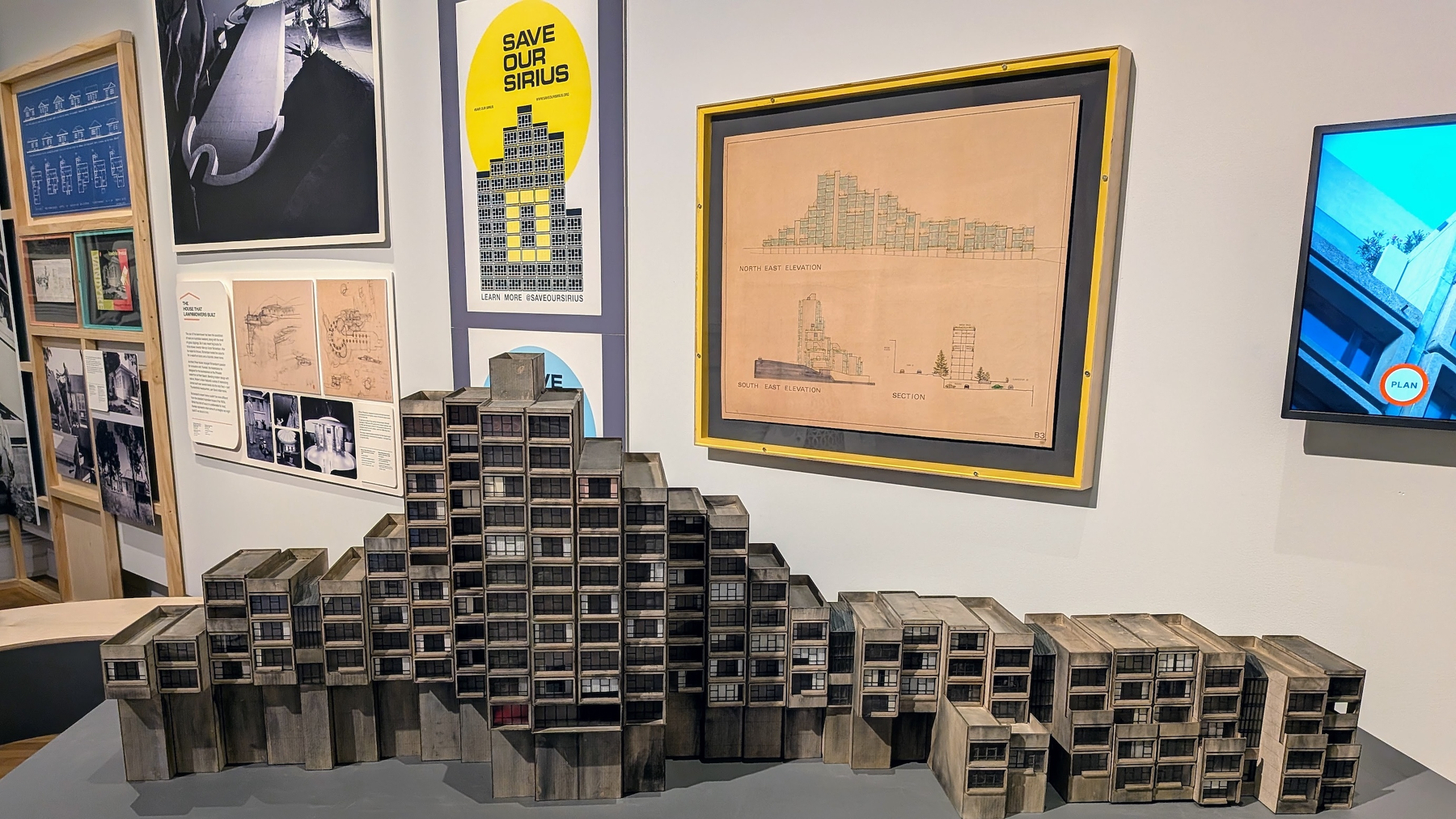 A detailed architectural model of Sydney&rsquo;s brutalist Sirius Building is displayed in a gallery setting surrounded by various framed posters and plans related to the structure.