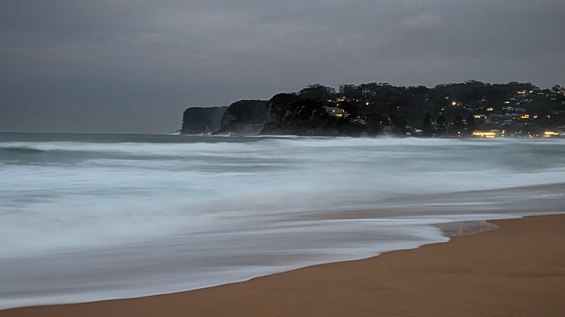 Waves wash onto a sandy beach under a cloudy sky at dusk, with a distant rocky headland and the scattered lights of houses clinging to the cliffs.