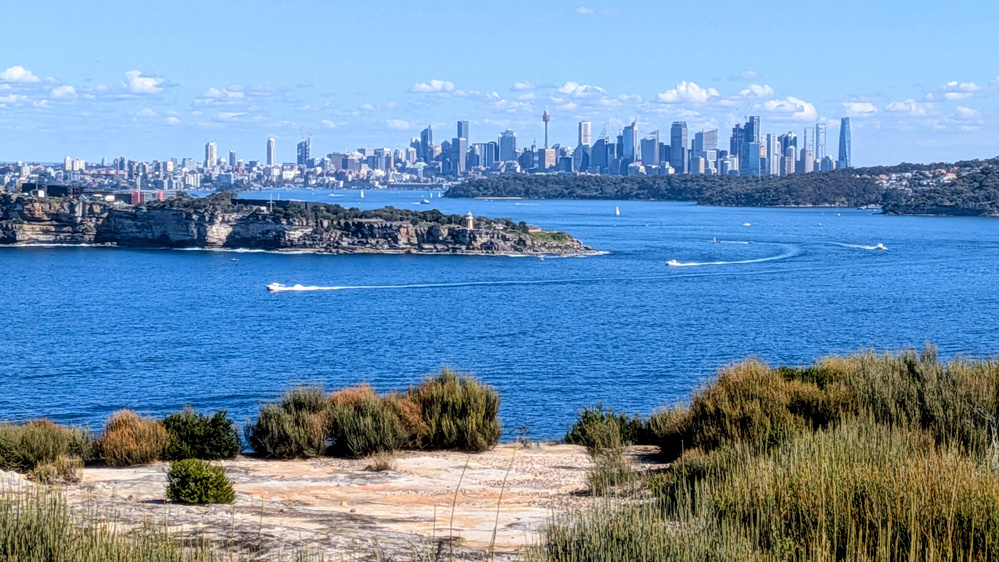 A coastal landscape at North Head, Sydney, features clear blue water with boats and a distant city skyline under a partly cloudy sky.