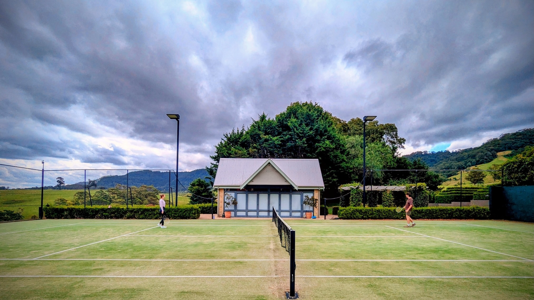 Two people are playing on an outdoor tennis court with forested hills and a cloudy sky in the background.