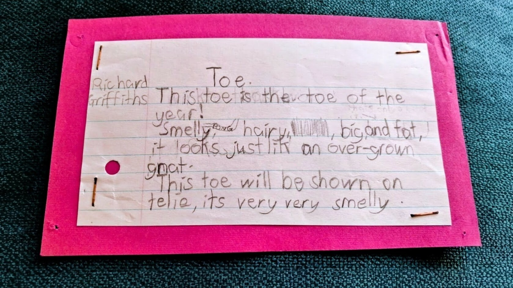 A handwritten note humorously describes a toe of the year with characteristics like being smelly, hairy, and big, written on lined paper stapled to a pink backing.