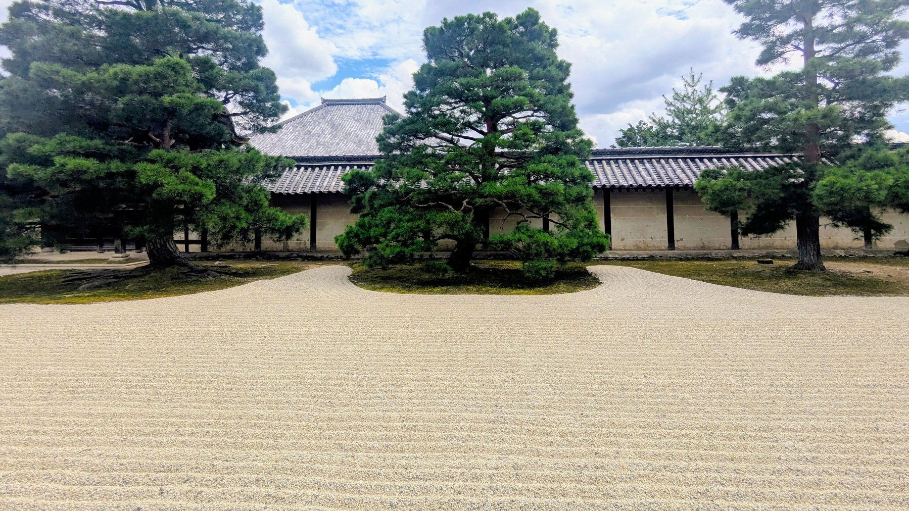 A traditional Japanese rock garden in Kyoto, featuring raked gravel with a few carefully placed pine trees stands in front of a traditional building with a tiled roof.