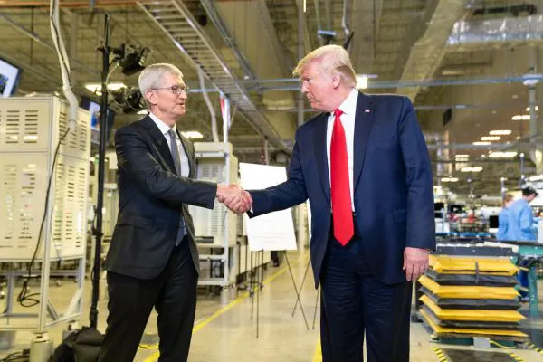 Two men are shaking hands in a factory setting with industrial equipment and workers in the background.