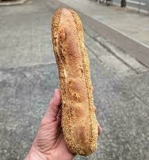 A person's hand is holding a baguette-like croquette on an outdoor pavement.