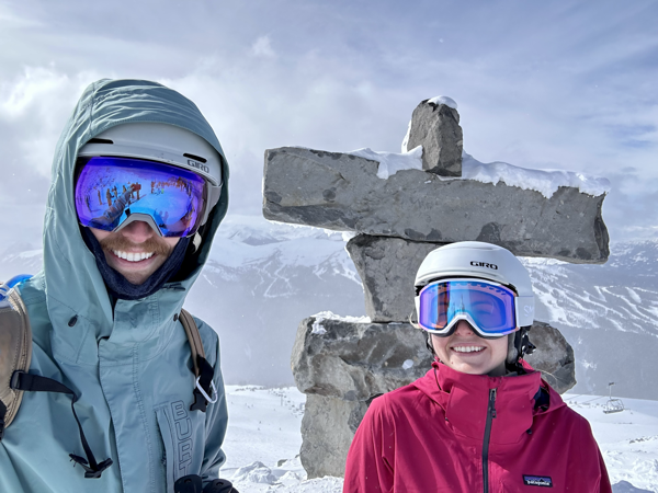 Catie and Nick, smile from the top of a snow mountain, in front of a large ~8ft stone man with mountains in the background