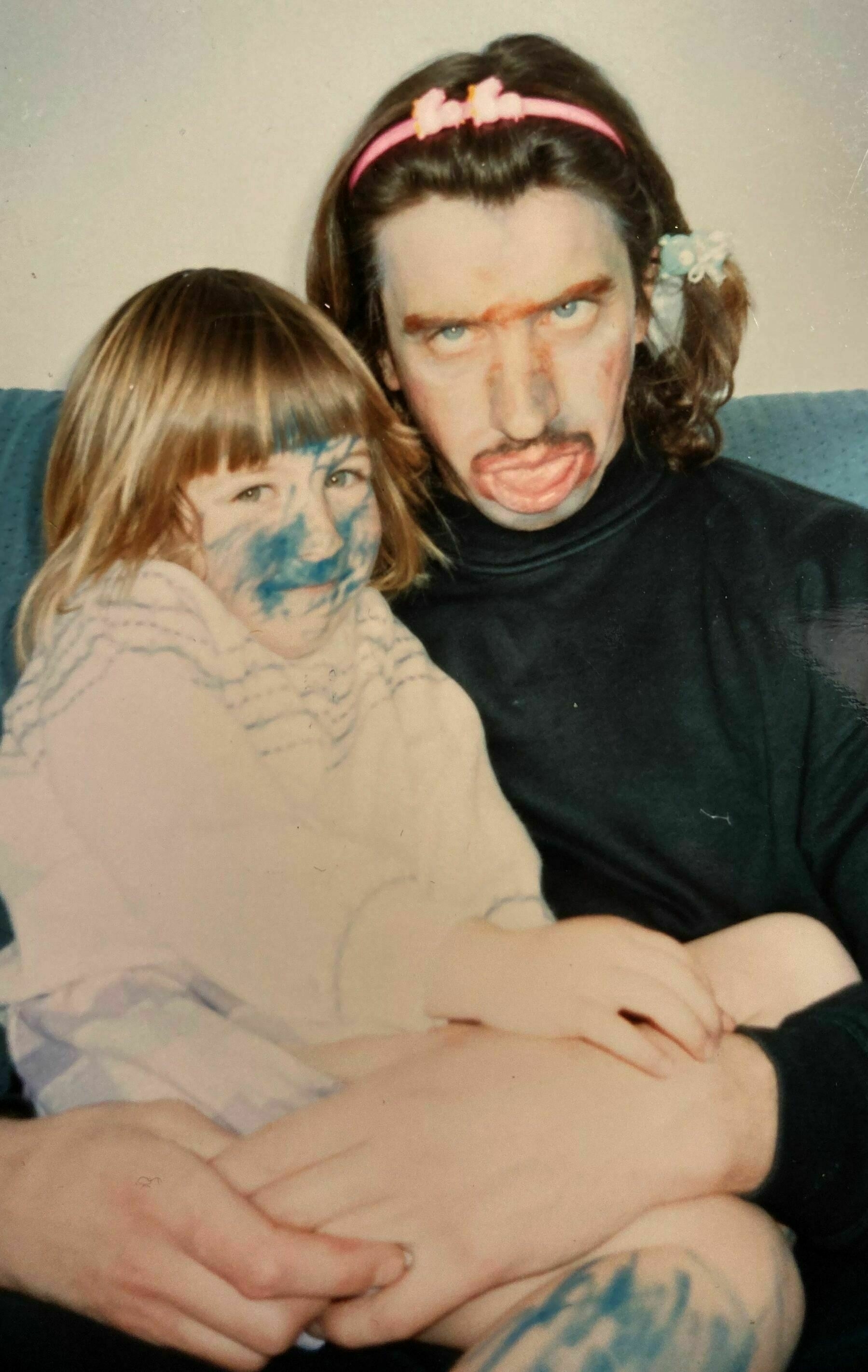 Man holding a child on his lap. The child has blue colouring on her face and leg the man has brown colouring on his face and an alice band in his hair.