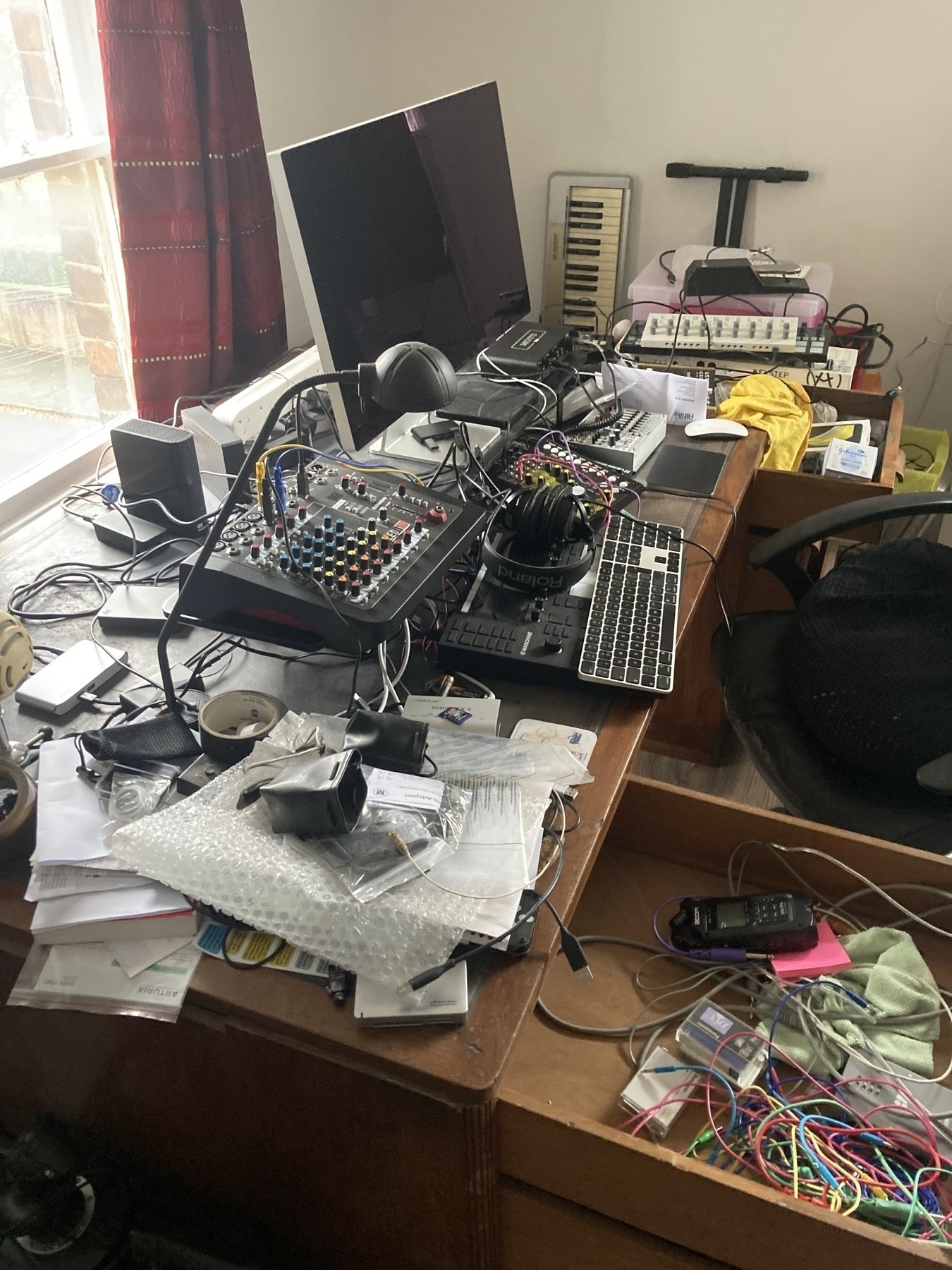 A cluttered desk is filled with audio equipment, cables, papers, and miscellaneous items.