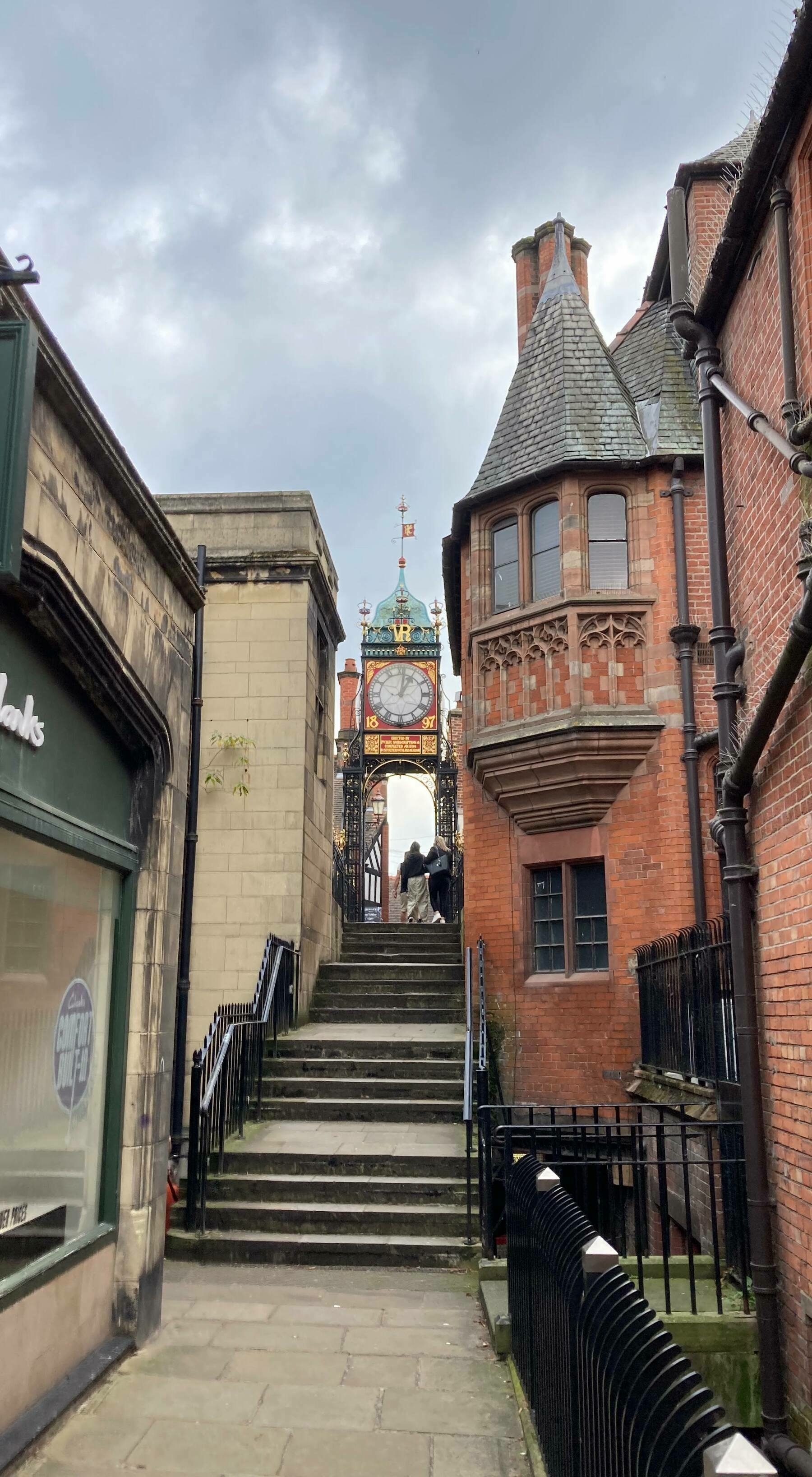 Paved walkway of Chester city wall passes between buildings as steps lead up to the Eastgate bridge which is surmounted by an ornamental clock dedicated to Queen Victoria.