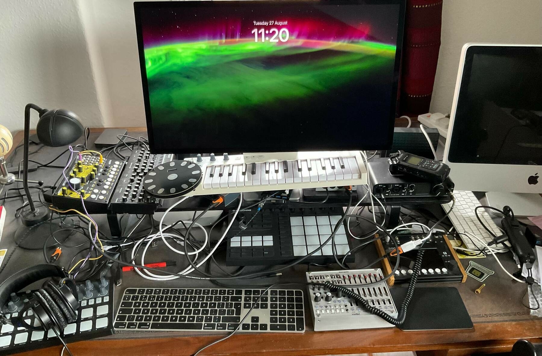 A cluttered desktop with two computers and their keyboards, a musical keyboard, several sequencers, synthesisers with a tangle of cables connecting some of them.   