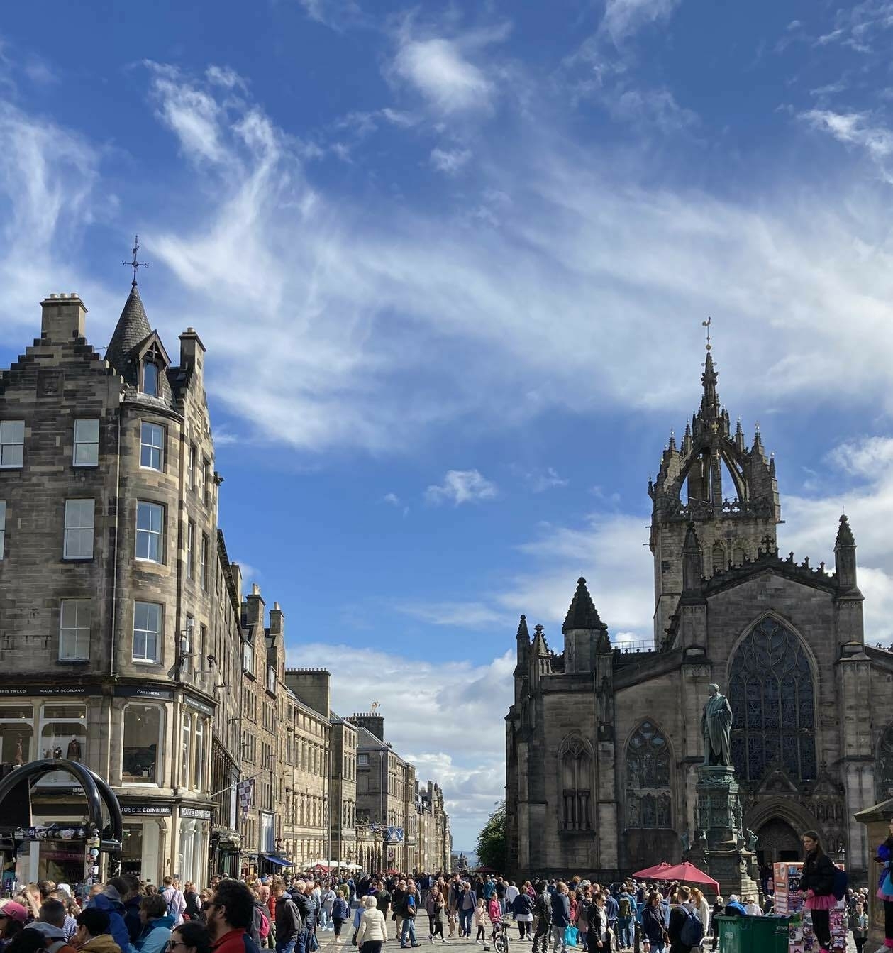 A view along the Royal Mile in Edinburgh. Saint Giles cathedral on the right and on the left a line of shops stretching into the distance most with five floors of residential apartments above them culminating in arrays of chimney stacks.