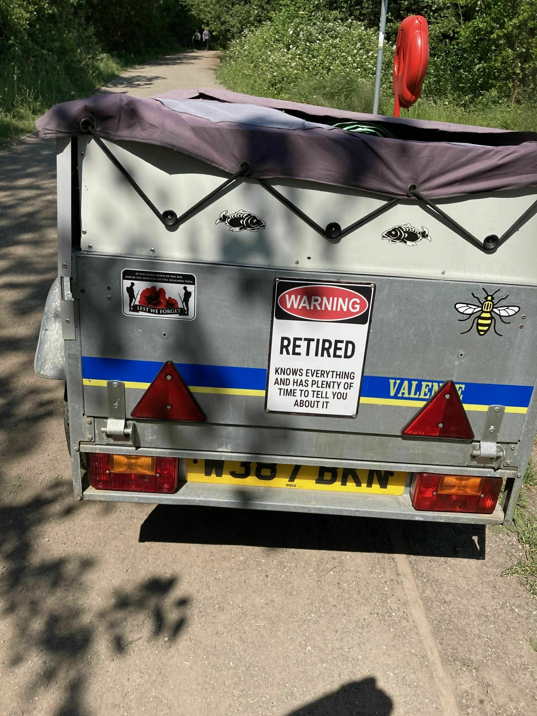  a large trailer containg fishing equipment beneath the covers.  On the back a large sign which reads "Warning. Retired.  Knows everything and has plenty of time to tell you about it."   