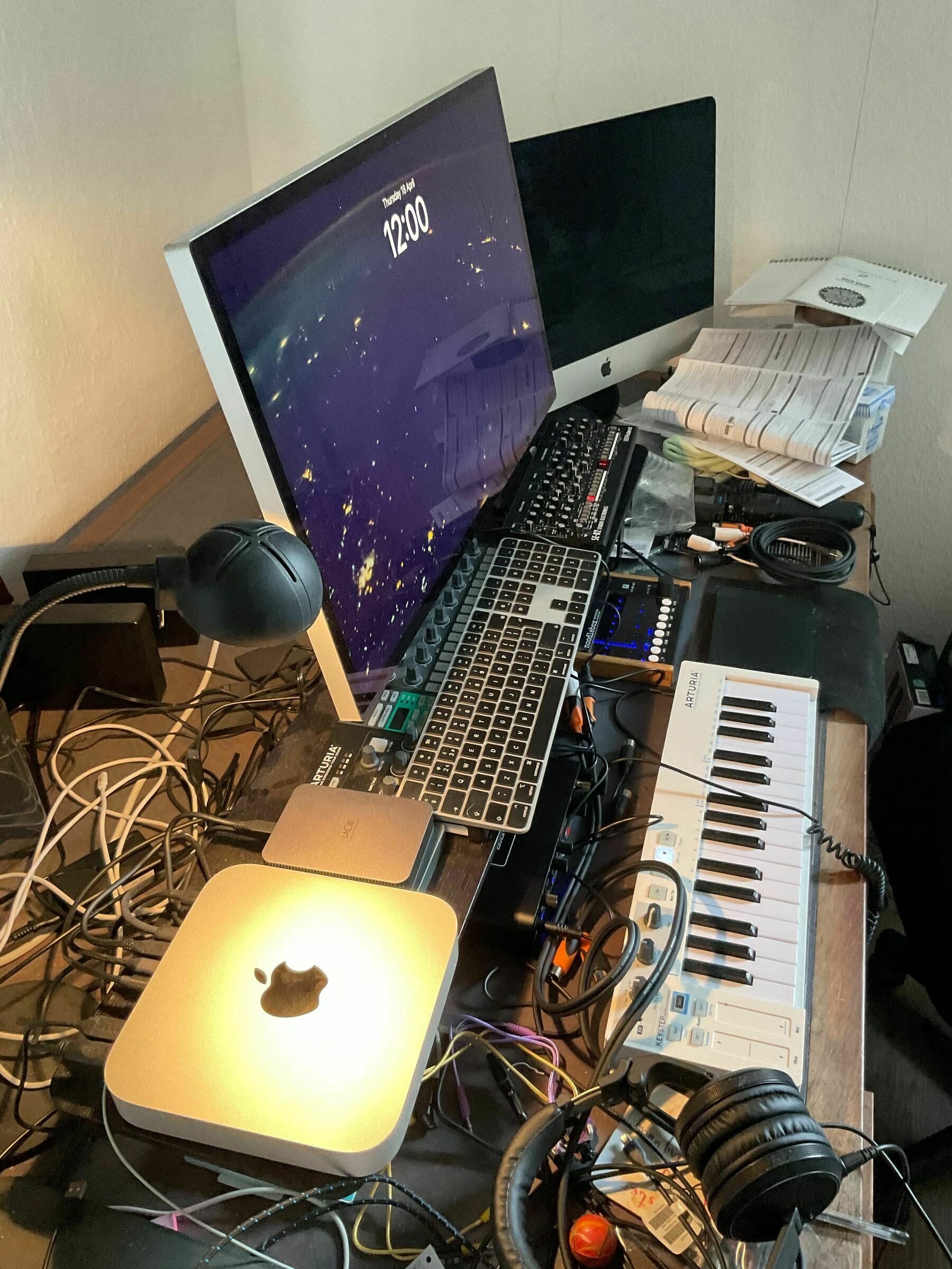  A cluttered desk with two computers, a tangle of wiring, musical keyboards and devices etc.