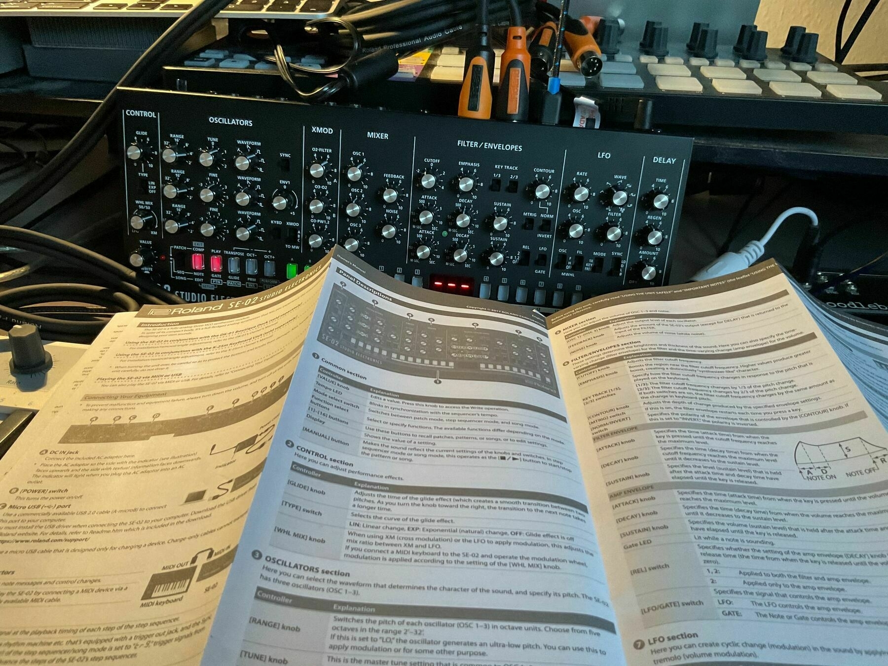 A cluttered desk of controllers and wires connected to a SE02 synth with the paper instruction manual in the foreground. 