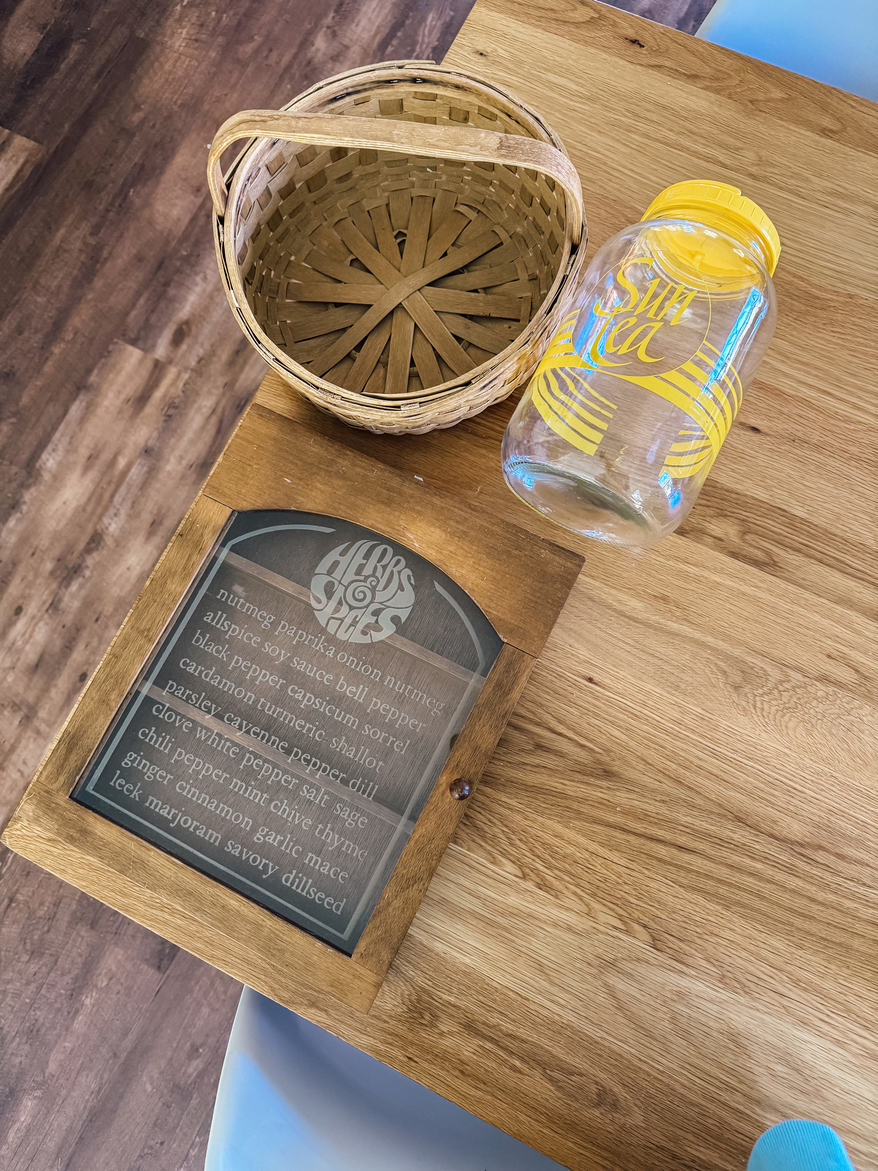 A wooden table holds an empty glass jar, a small woven basket, and a wooden herb and spice cabinet.