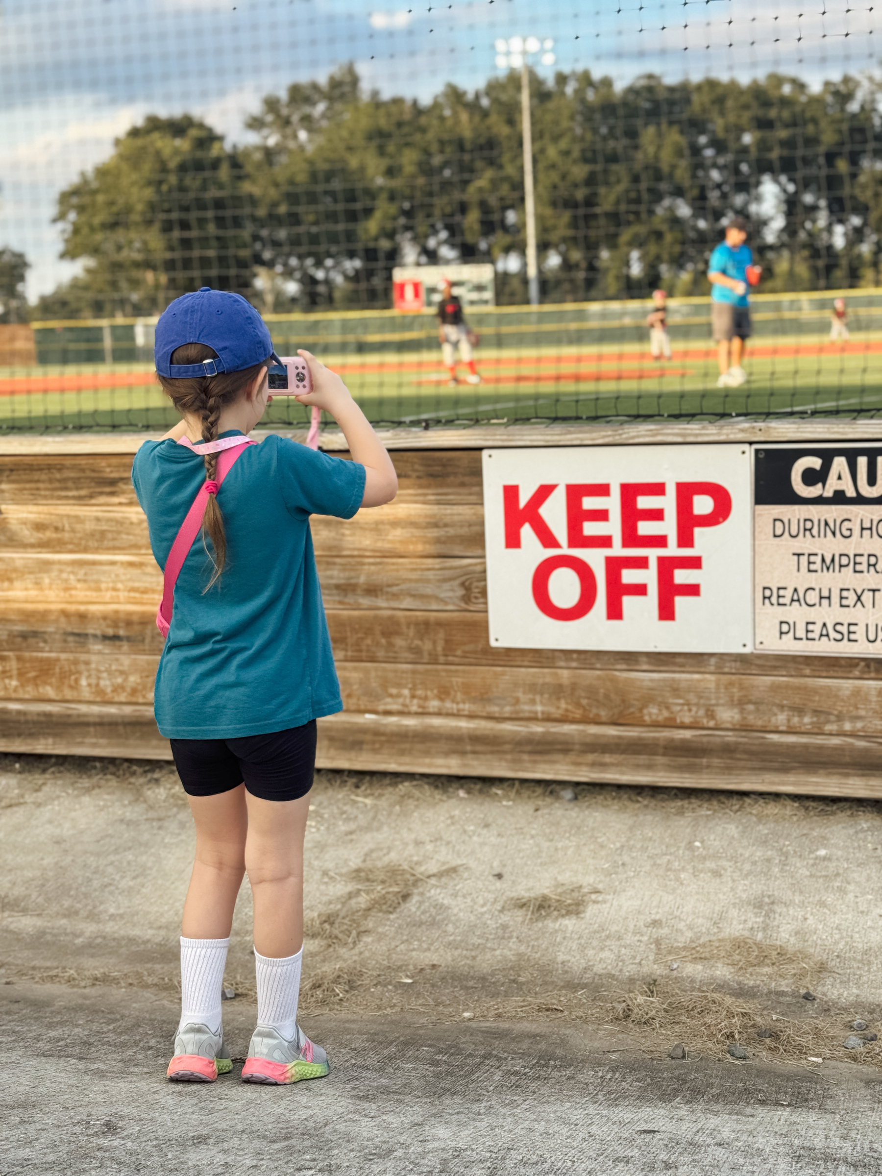 A child in a blue shirt and hat takes a picture through a netting, while a "KEEP OFF" sign is visible in the foreground and a baseball field is in the background.