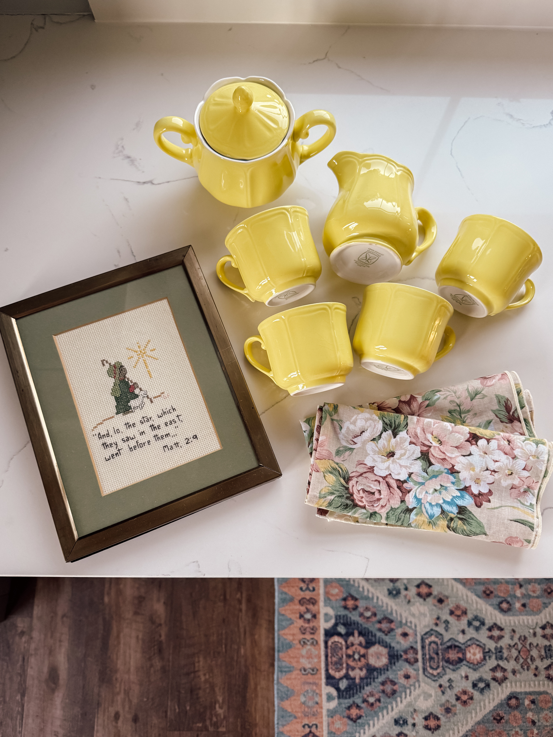 A set of yellow dishes, along with a framed cross-stitch and floral napkins, are arranged on a countertop next.