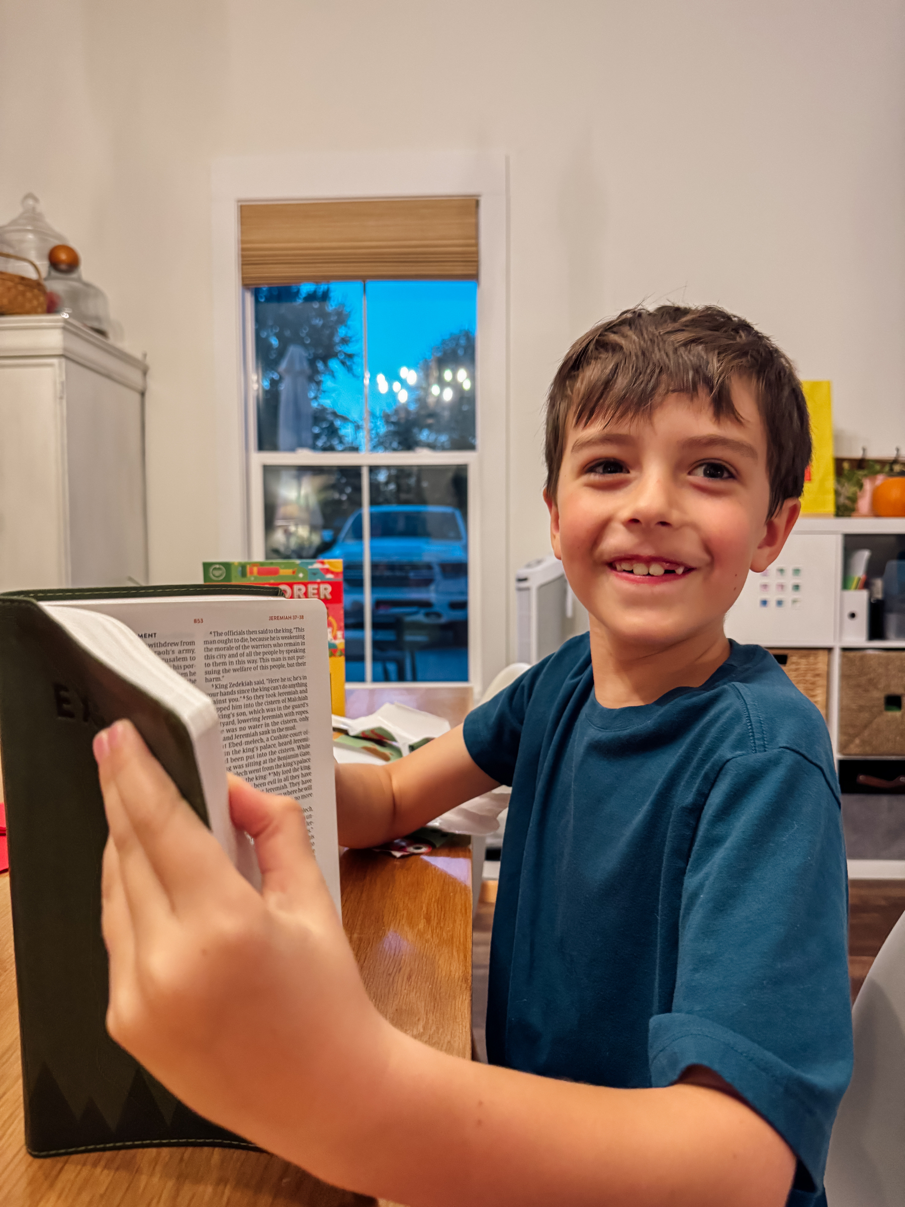 A smiling child in a blue shirt holds an open Bible while sitting at a table.