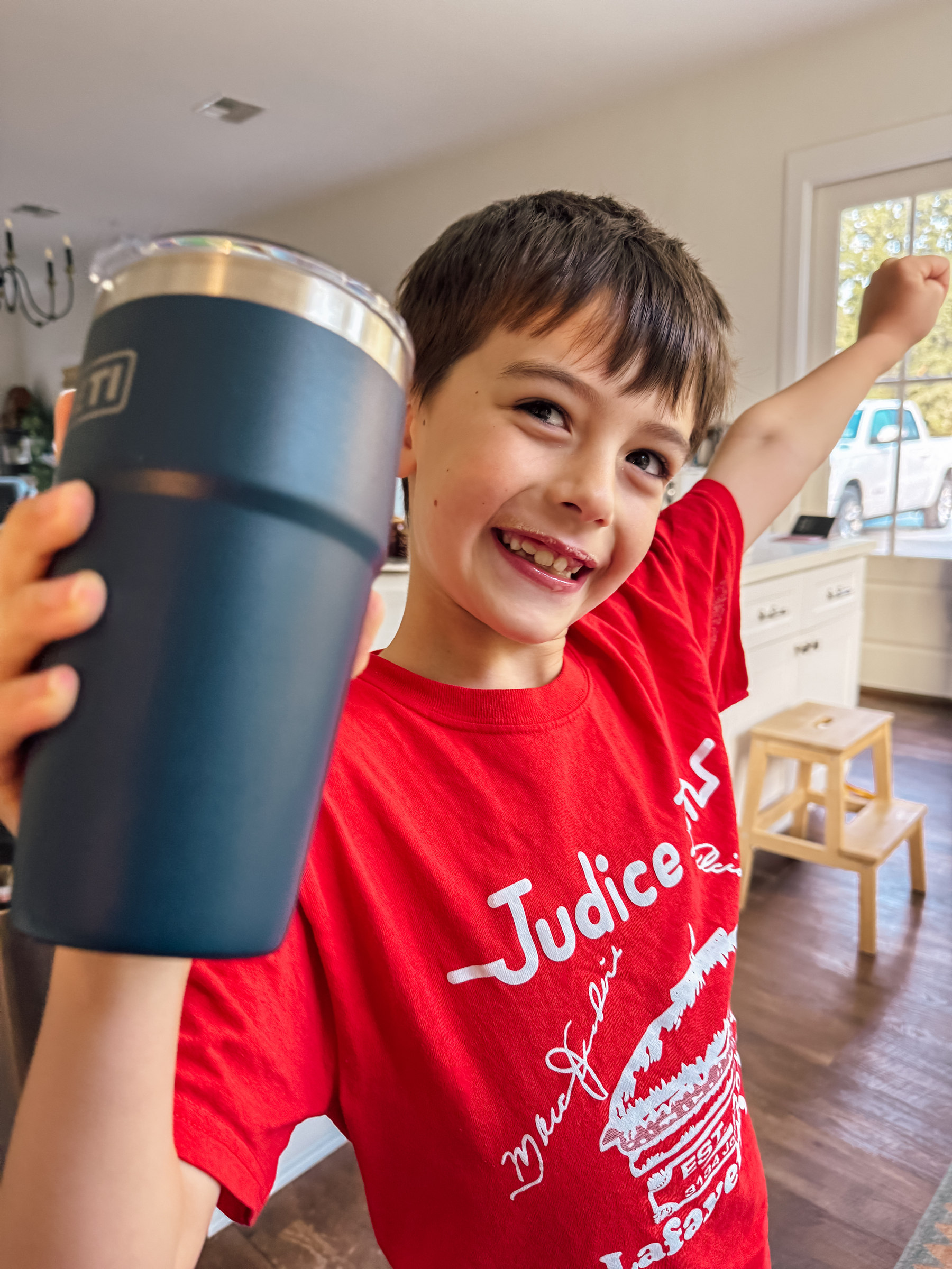 A smiling child wearing a red t-shirt is holding up a blue tumbler in a kitchen setting.