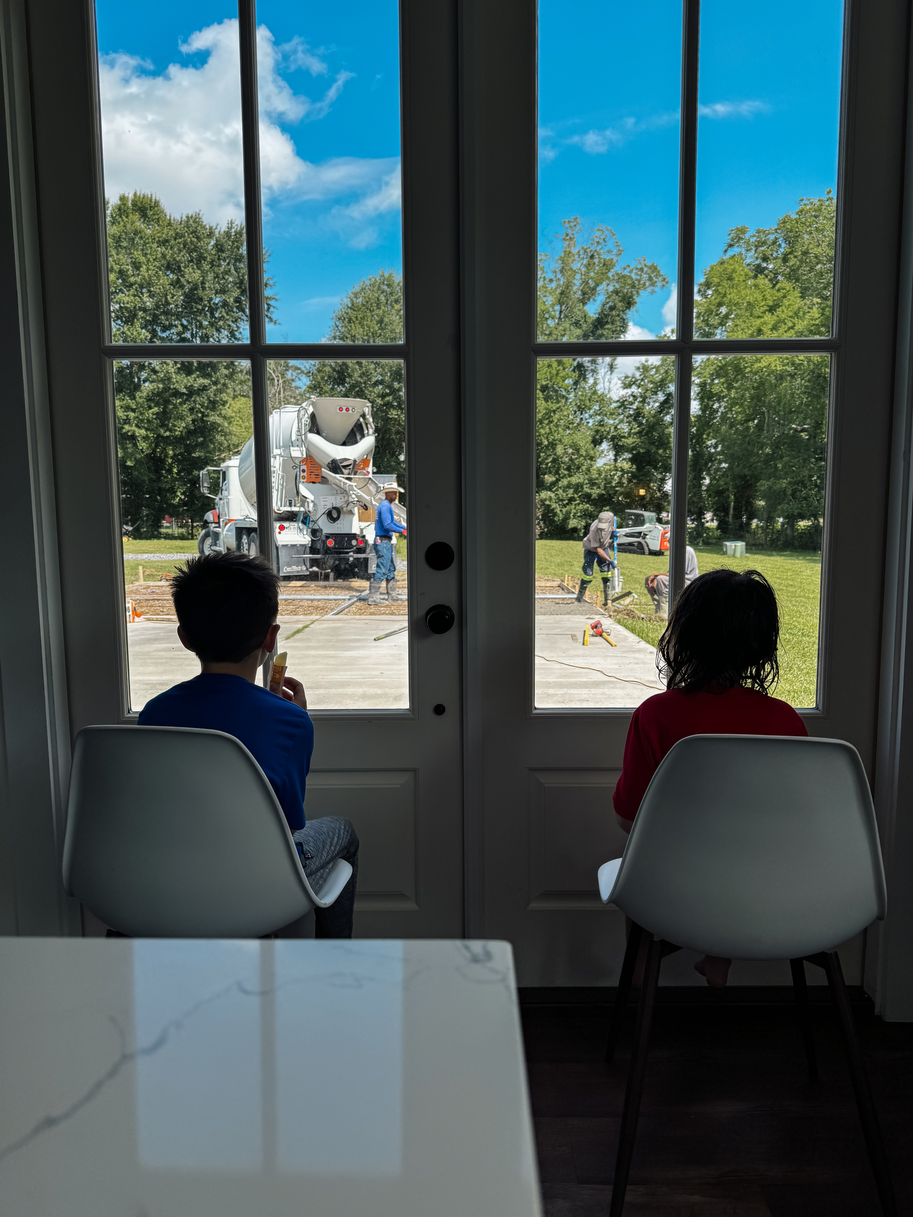 Two kids sitting in chairs in front of a window watching a concrete mixer pour concrete and men work to make it a slab for a porch
