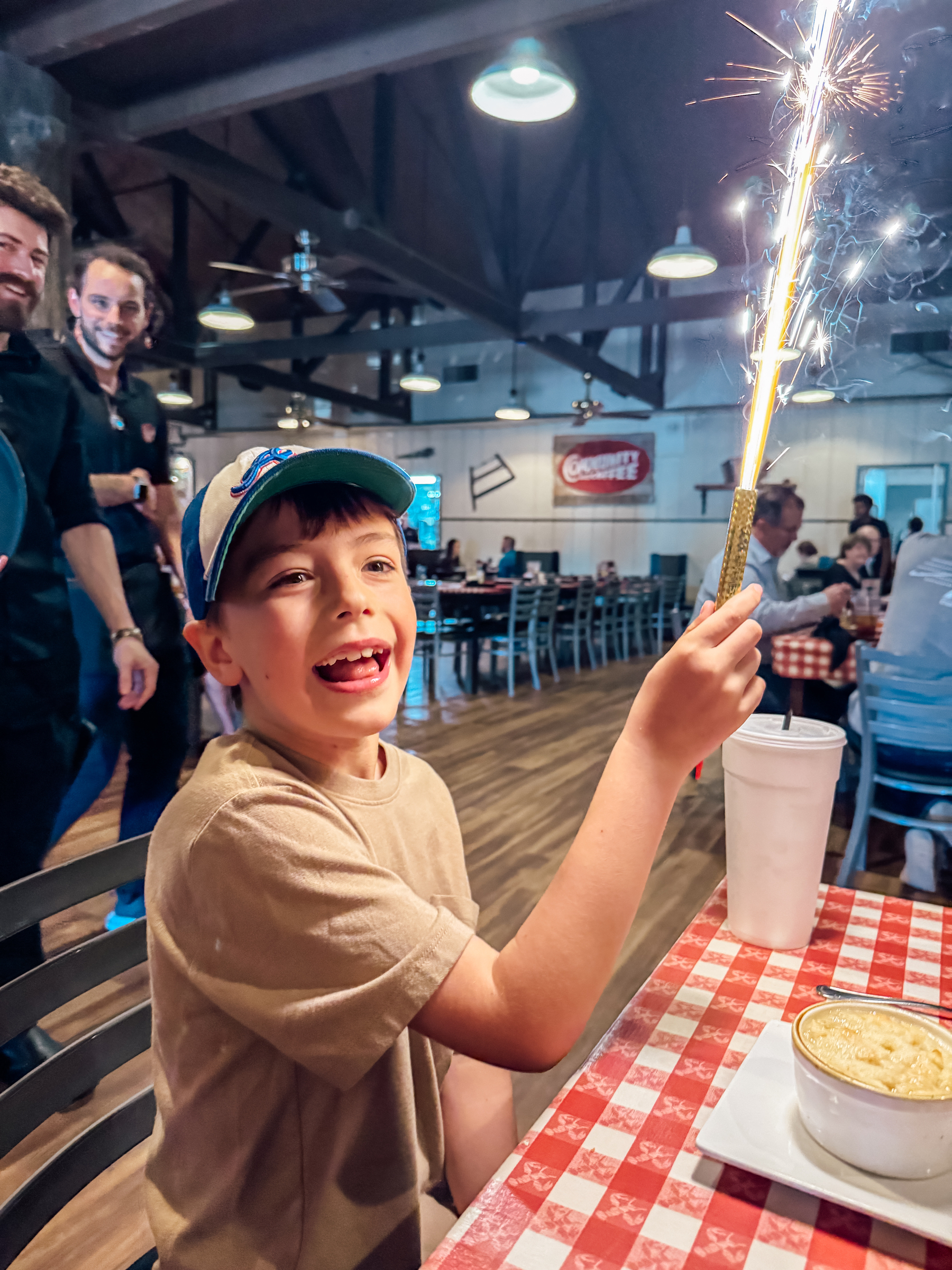 A child wearing a cap joyfully holds a lit sparkler while sitting in a dining setting with other people around.