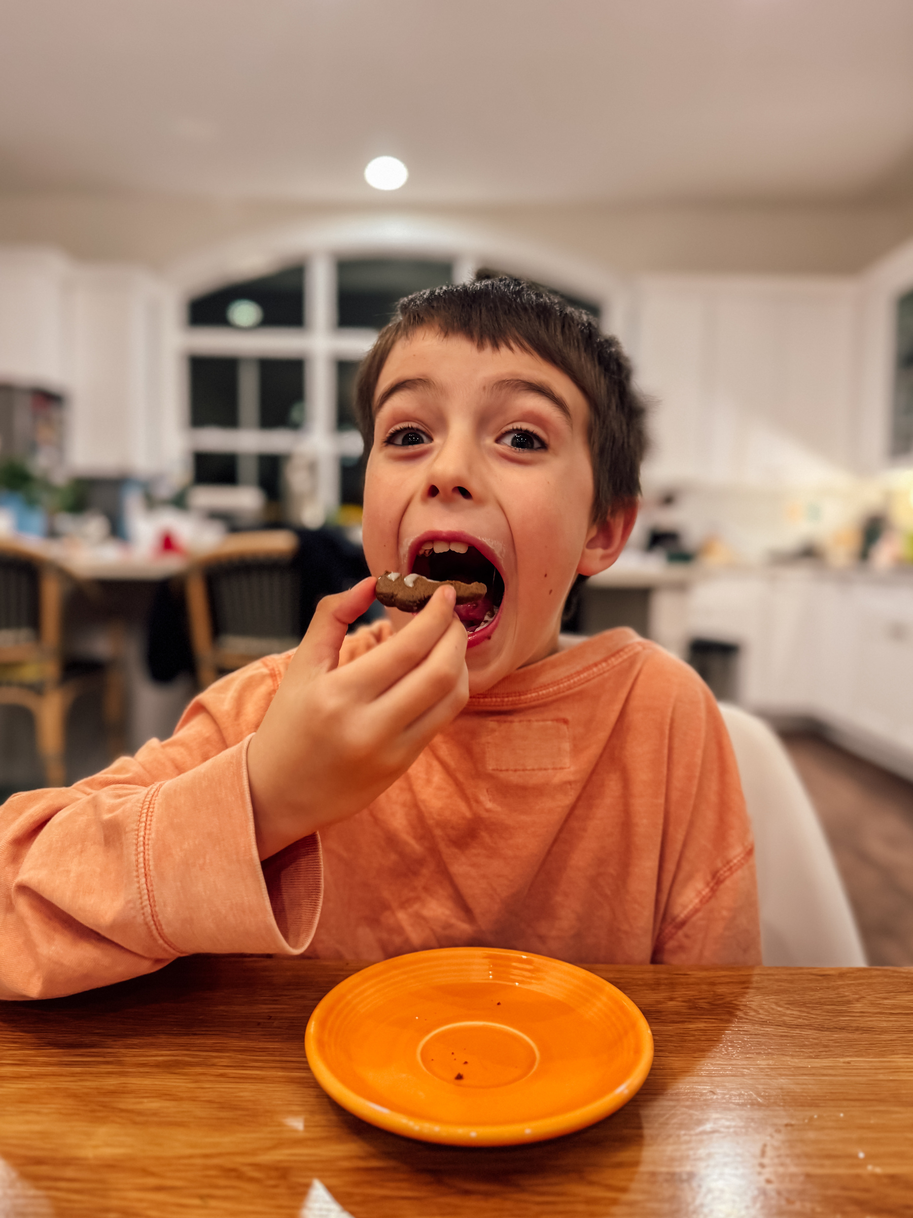 A child wearing an orange shirt is excitedly eating a cookie at a table with a small orange plate.