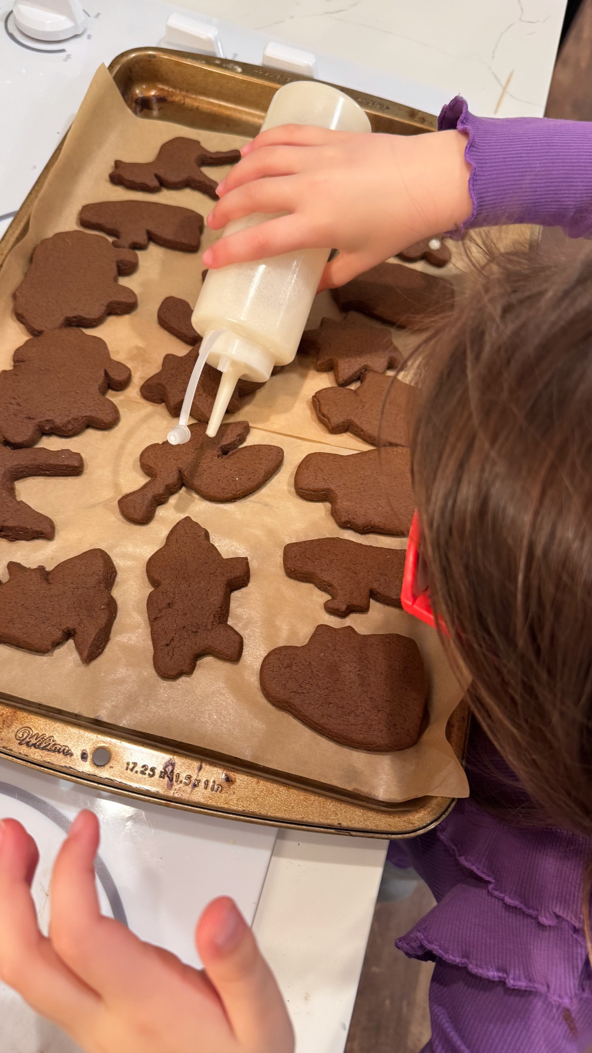 A child is decorating freshly baked gingerbread cookies with white icing on a parchment-lined baking sheet.