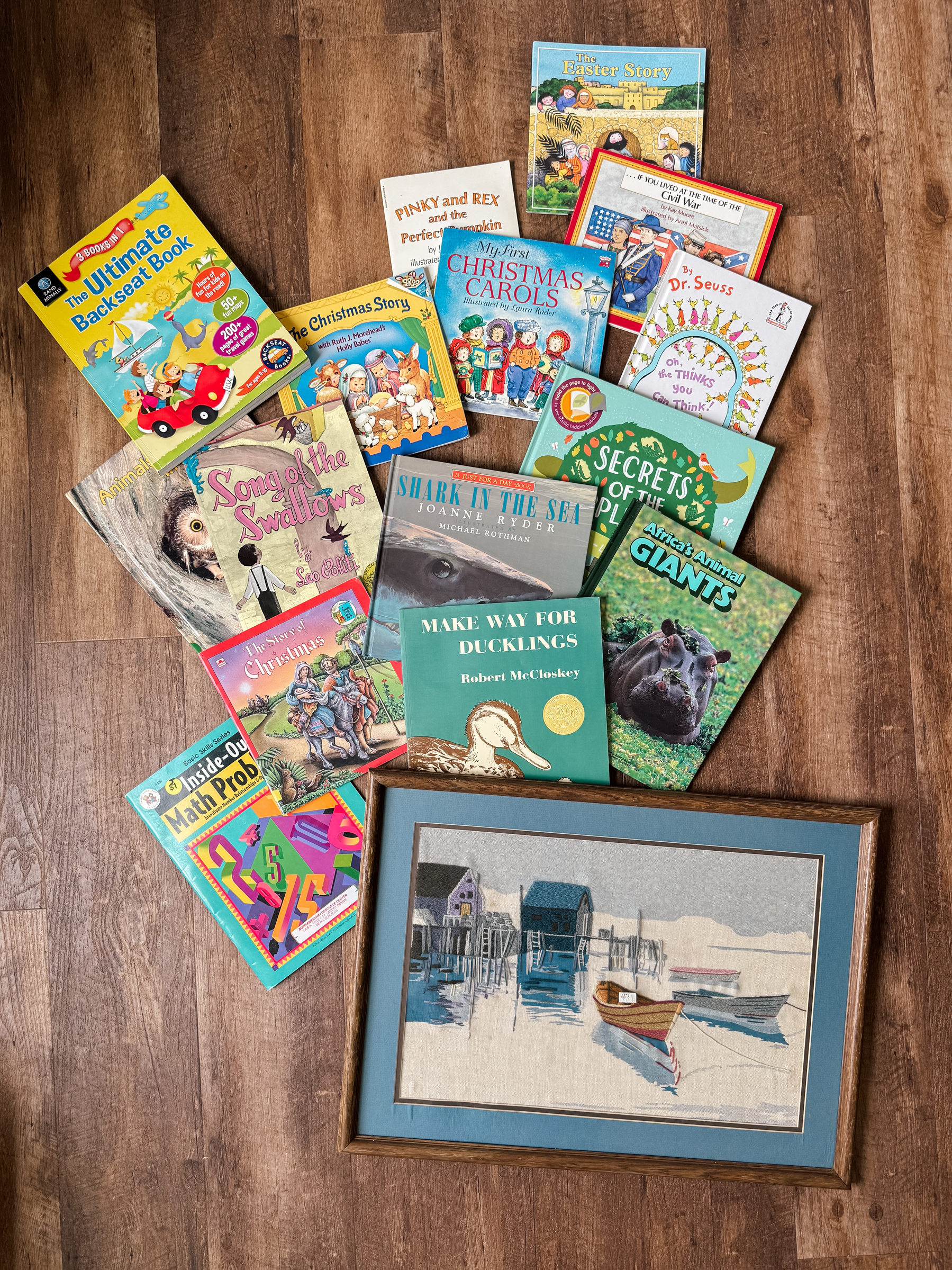 A photo of 15 children’s books and a framed embroidered portrait of some canoes along a pier