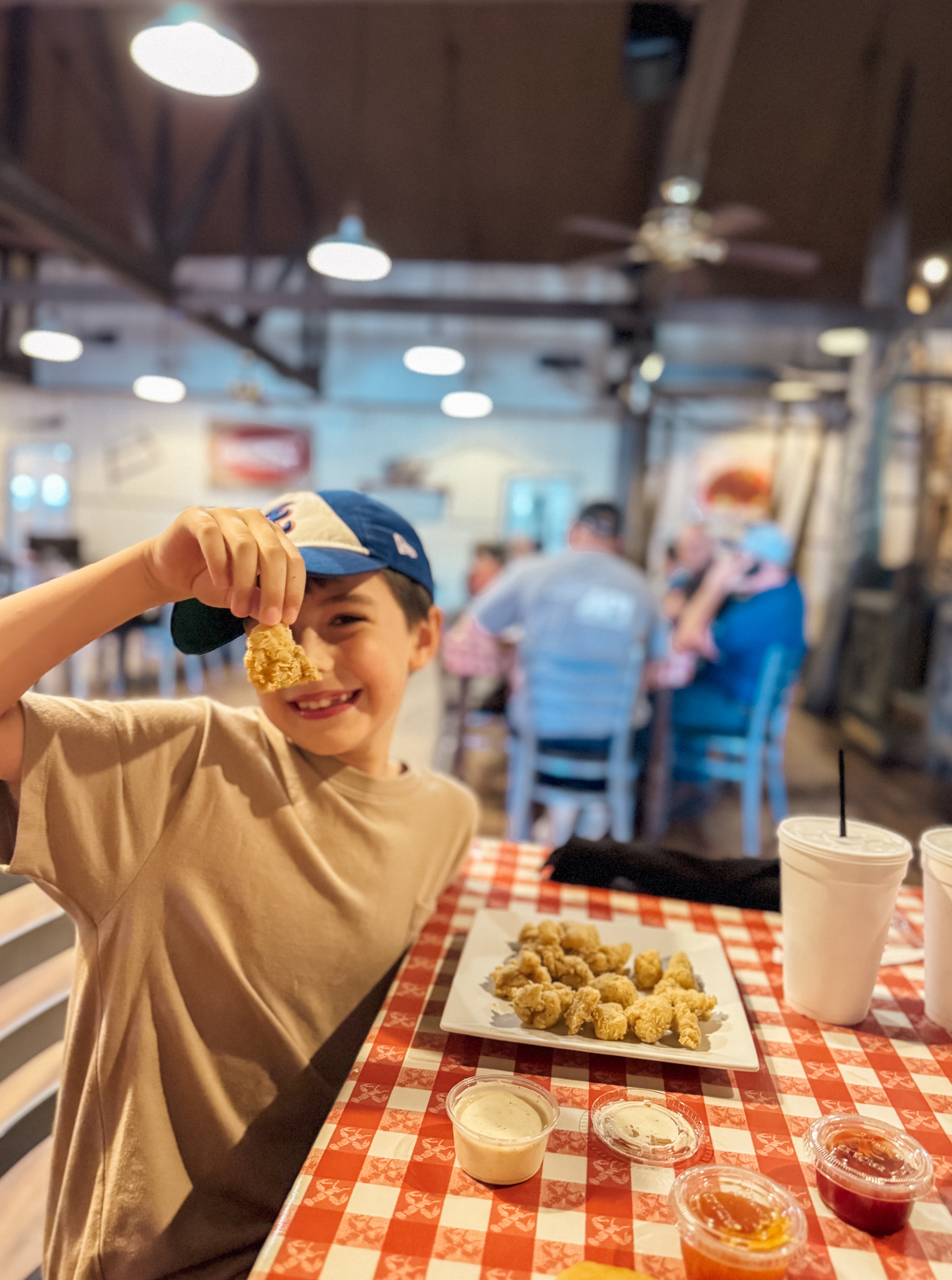 A smiling child is holding up a piece of fried alligator while seated at a table with a checkered tablecloth and dishes of food and sauces in a restaurant setting.