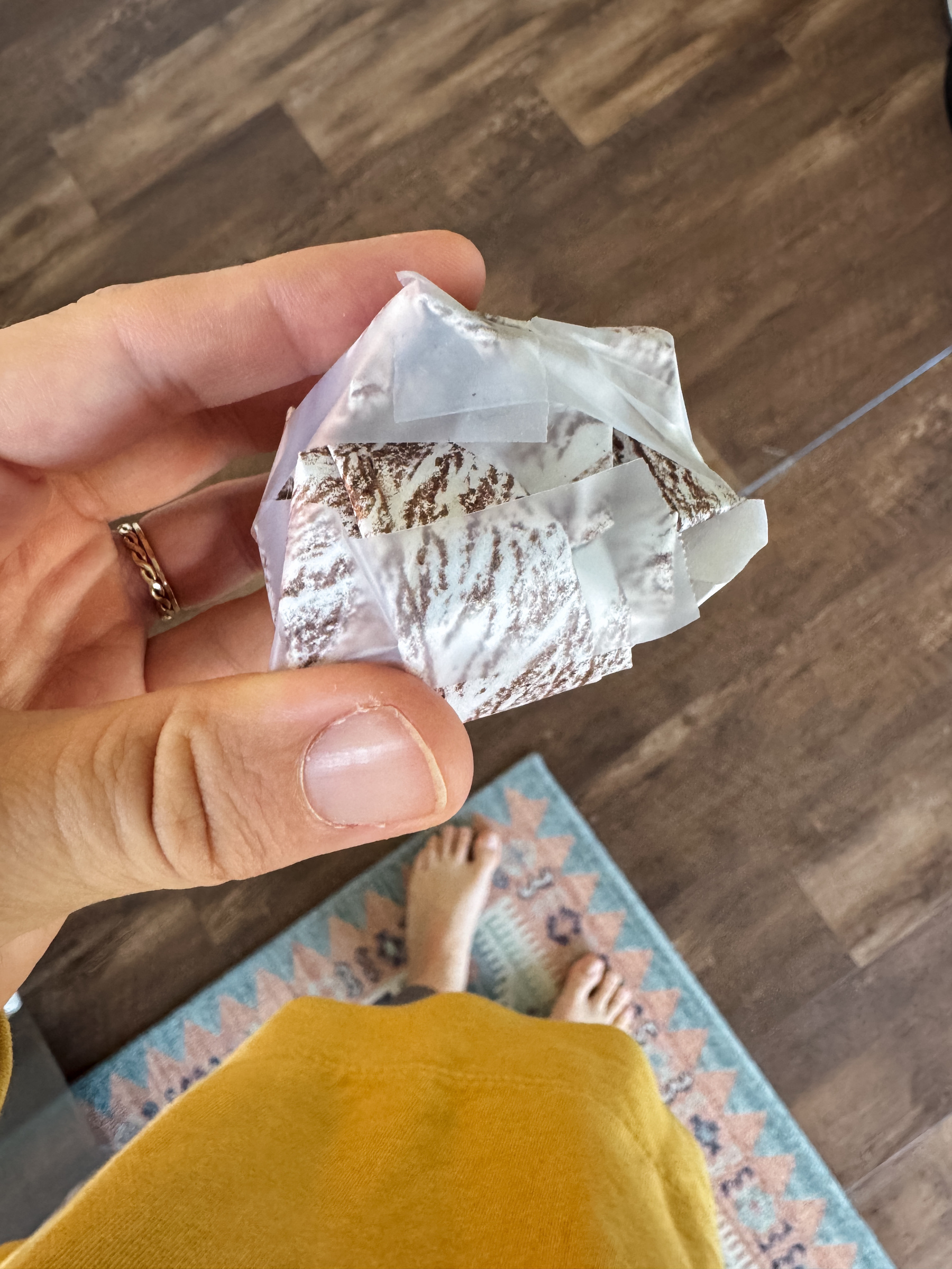 A person holds a crumpled and taped piece of paper colored brown and meant to be a cannonball above a patterned rug on a wooden floor.