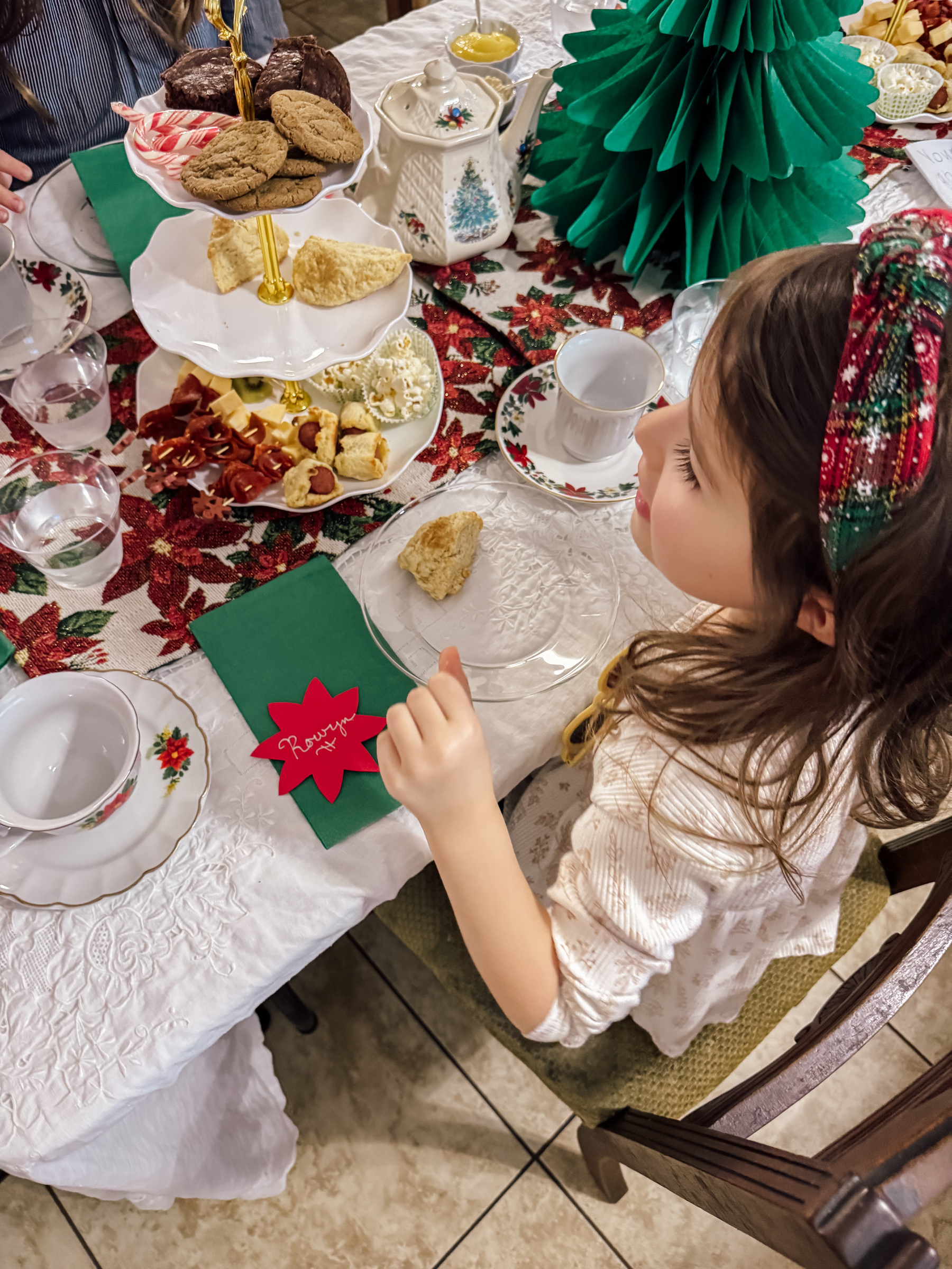 A young girl sits at a festively decorated table with holiday treats, a Christmas tree centerpiece, and empty teacups awaiting tea.