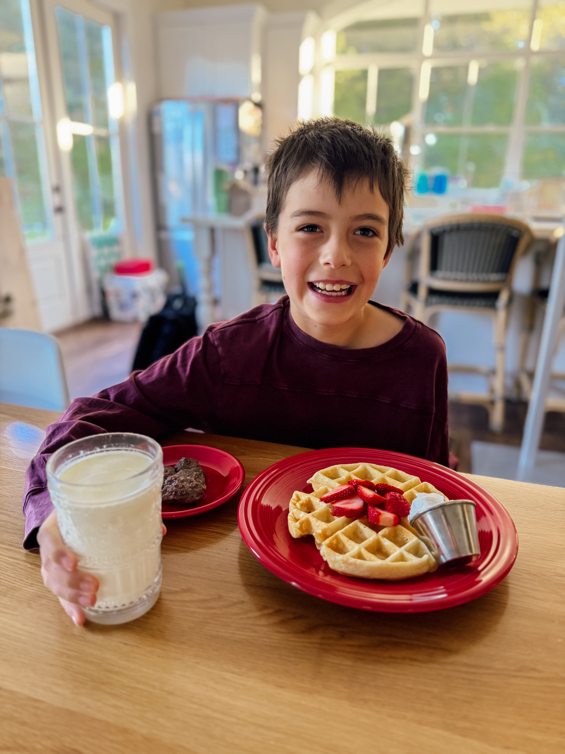 A smiling child sits at a table enjoying a breakfast of waffles with strawberries, milk, and sausage.