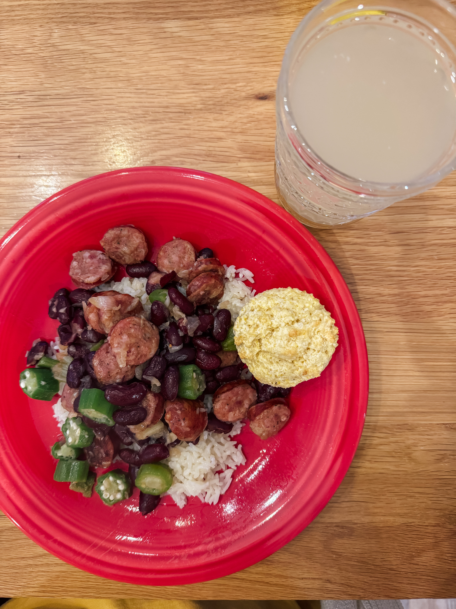 A red plate with rice, sausage, beans, and sliced okra is paired with a small cornbread muffin, next to a glass of light-colored beverage.