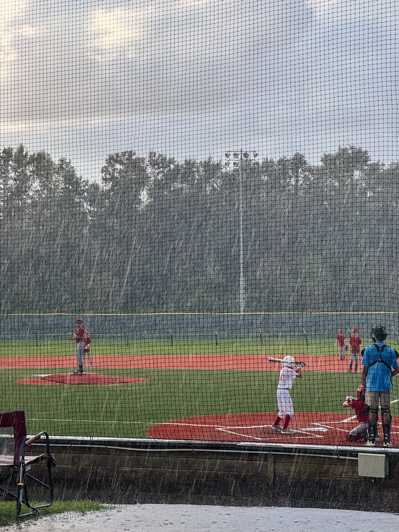 Auto-generated description: A baseball game is taking place in the rain on a field surrounded by trees, with one player pitching and another at bat.